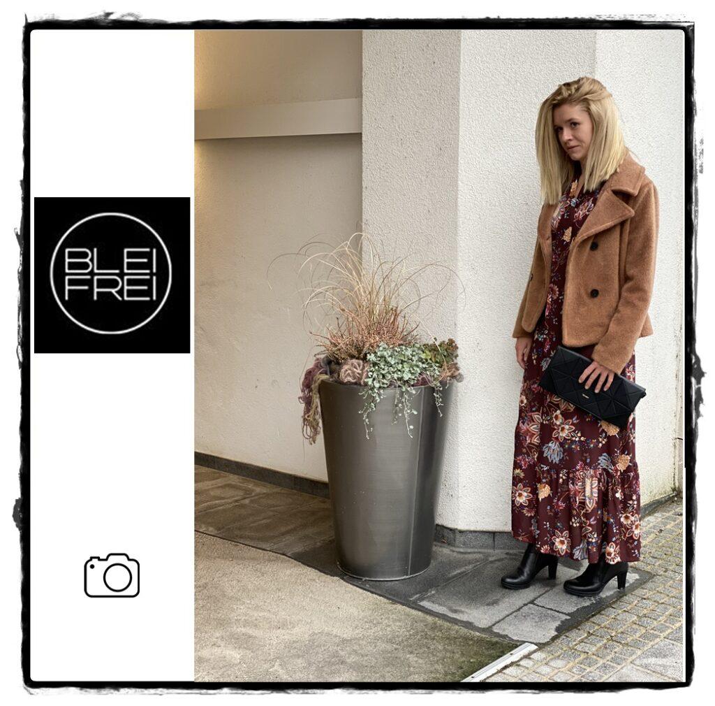 Frau steht neben einem großen Blumenpot mit einer roten Blume, trägt einen braunen Mantel und ein florales Kleid